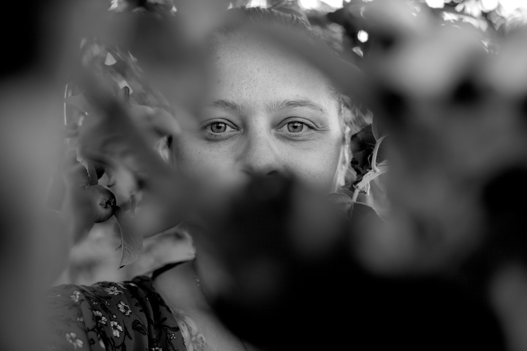 Portrait en noir et blanc d'une femme montrant son visage à travers des branches, avec un regard chaleureux et authentique.
