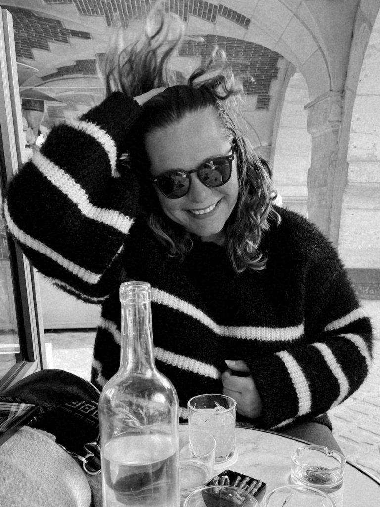 Une femme souriante avec des lunettes de soleil, portant un pull noir avec des rayures blanches, assise à une table avec des verres et une carafe d'eau, en train de jouer avec ses cheveux dans un cadre convivial.