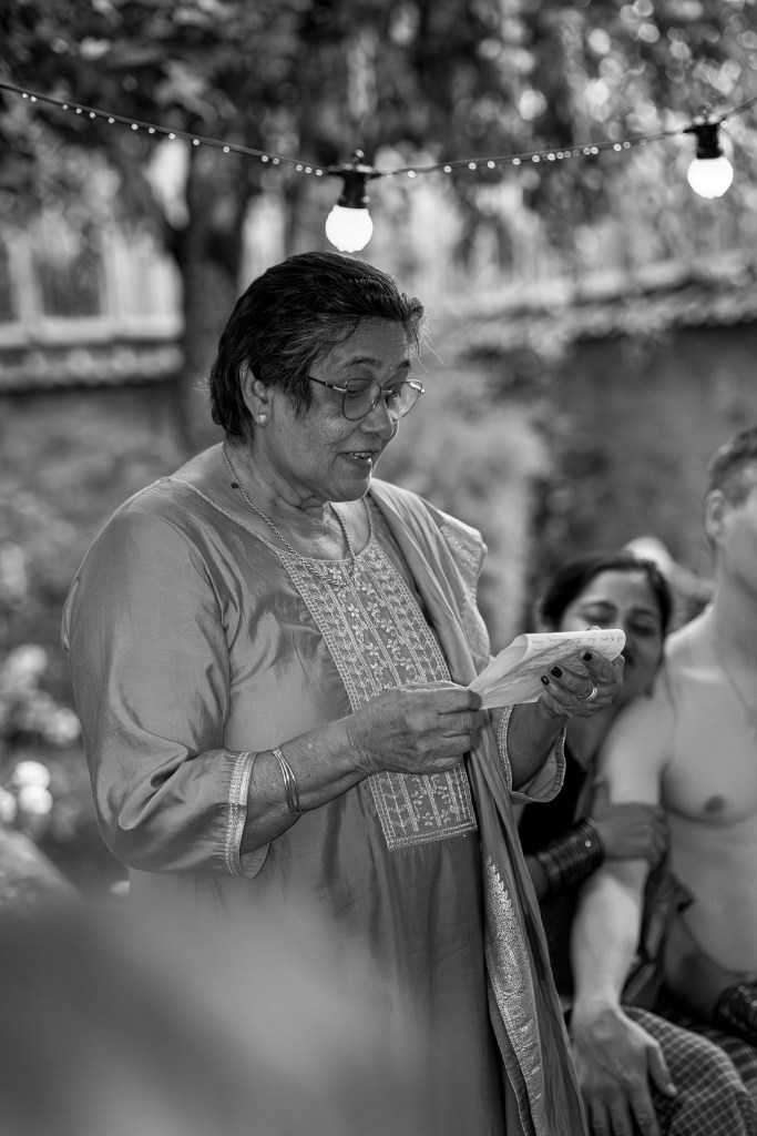 Une femme en tenue traditionnelle, lisant un texte lors d'une cérémonie, entourée de participants attentifs dans un jardin décoré de lumières.